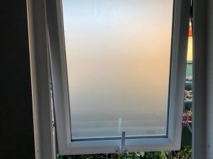 Modern awning window with frosted glass partially open, showing plants outside and minimal light filtering through.