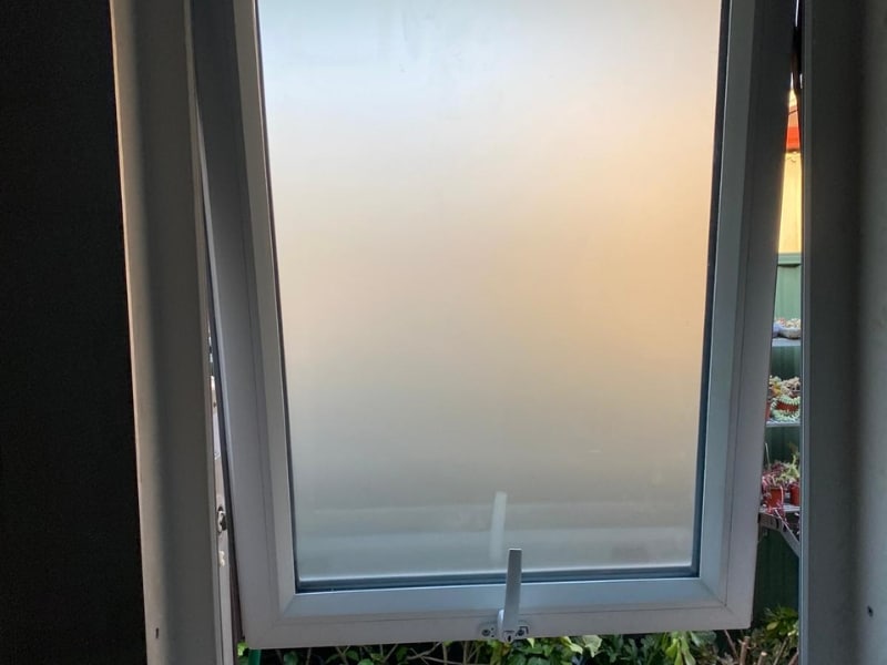 Modern awning window with frosted glass partially open, showing plants outside and minimal light filtering through.