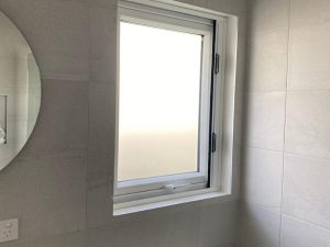 Modern awning window in a minimalist bathroom with frosted glass, tiled walls, and a circular mirror on the side.