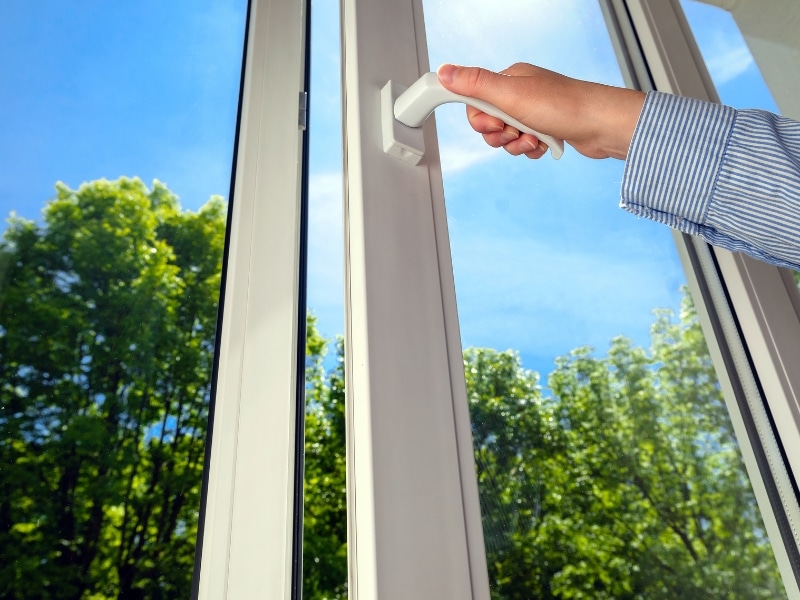 A person's hand is shown opening white upvc tilt turn windows, with a view of green trees and a blue sky outside.